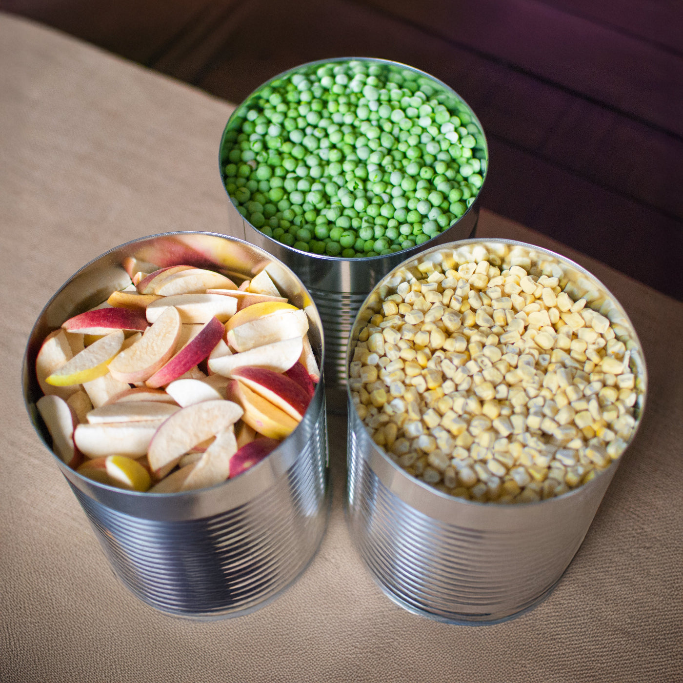 Cans filled with green peas, dried fruits, and corn on a brown surface.