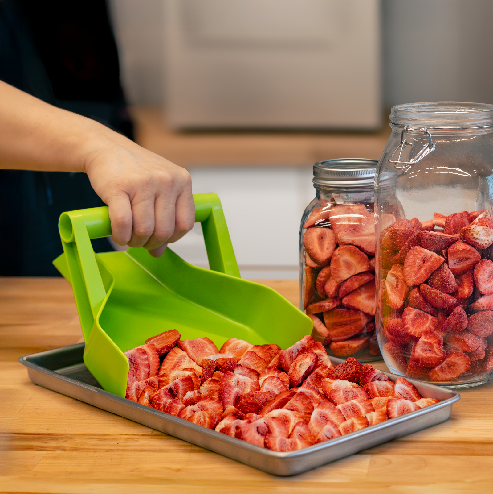 Person using a green silicone scoop to transfer dried strawberries from a tray into jars.