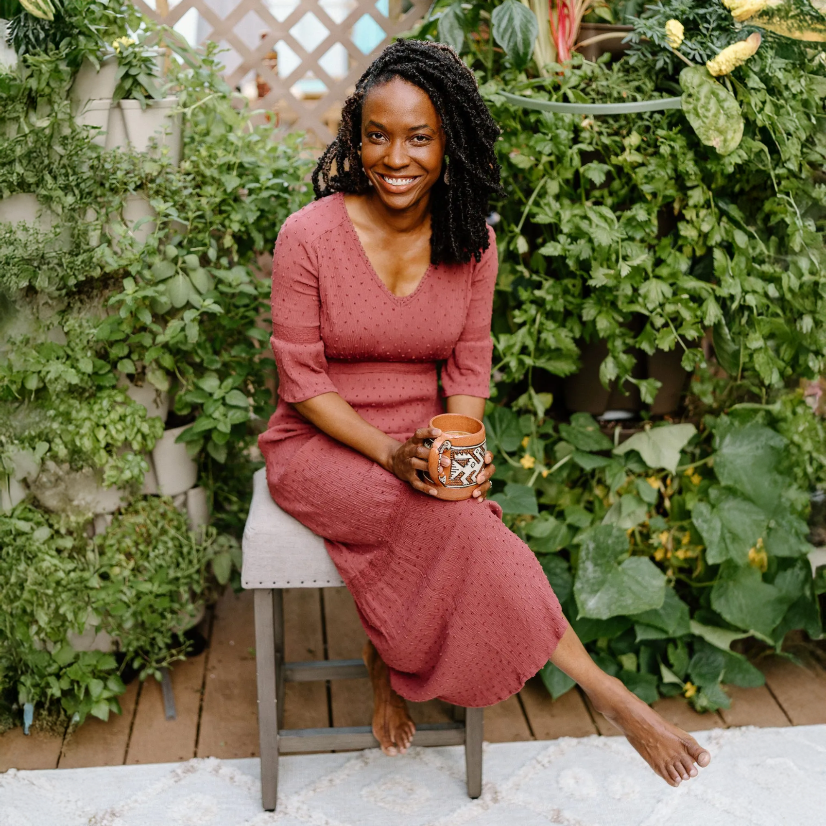 Woman in a pink dress sitting on a stool holding a cup in an outdoor garden setting.