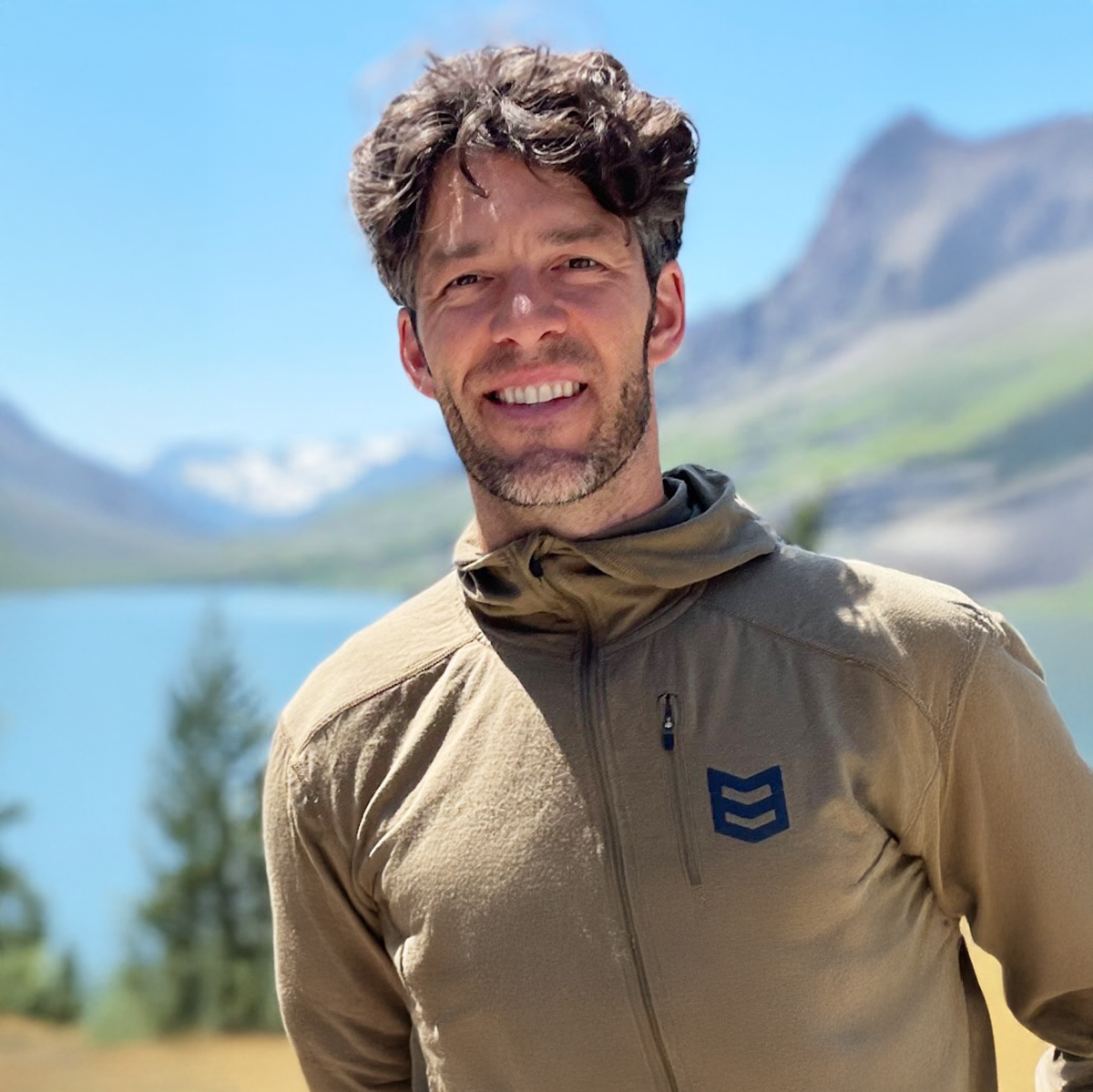 Man wearing a beige jacket with a logo in front of a mountainous landscape