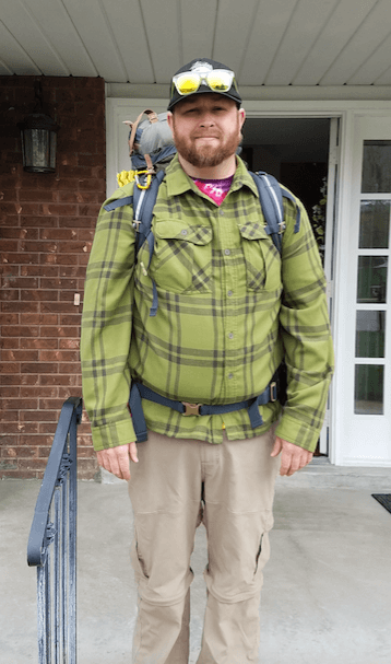 A man wearing a green plaid long sleeve shirt, khaki pants, a backpack, sunglasses and a hat