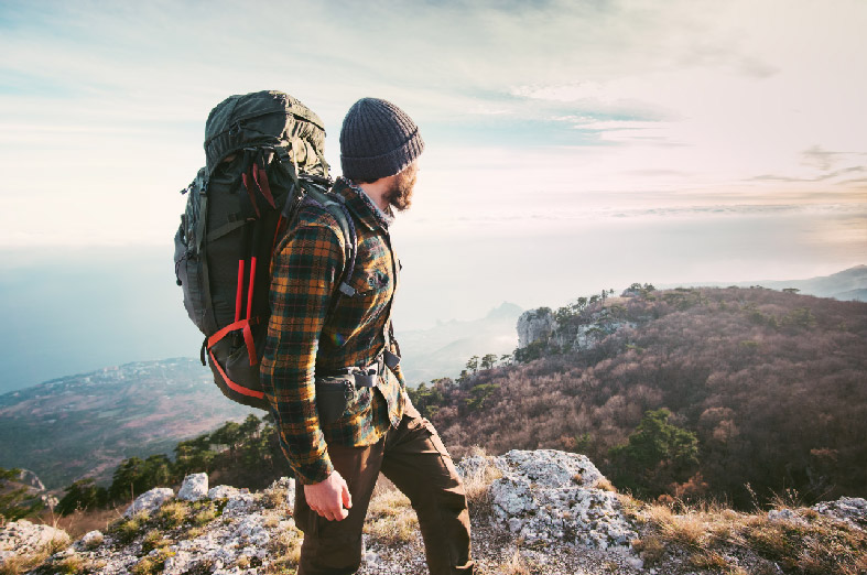 A man dressed in a beanie, a plaid long sleeve shirt, dark pants, and wearing a large hiking backpack, standing on a mountain.